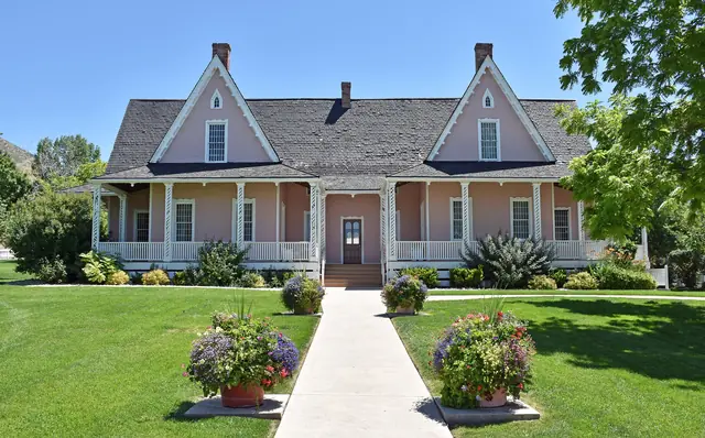 The pink Brigham Young Forest Farmhouse with a long porch, lawn and flower pots along the front walk